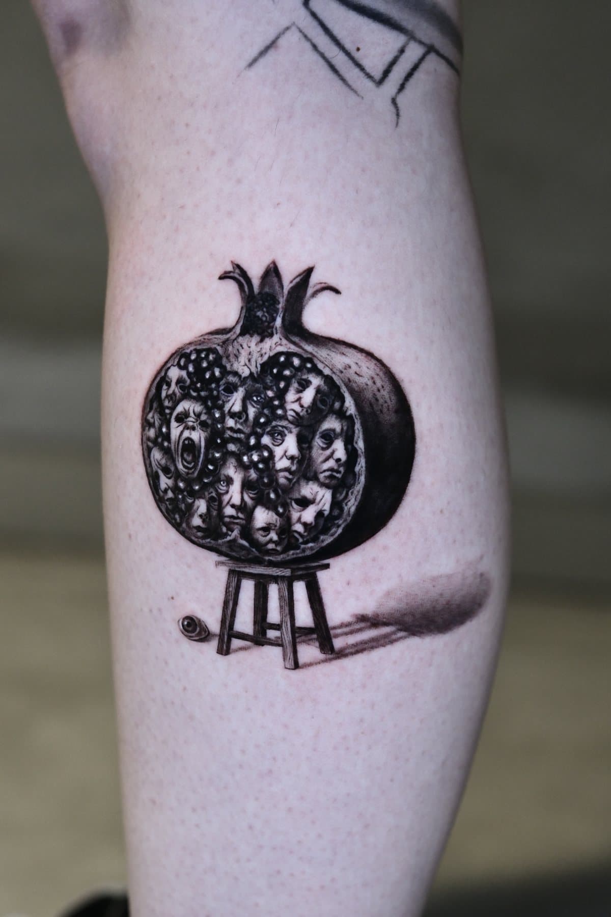 Black and grey surreal micro realism tattoo of a split pomegranate filled with human faces, resting on a small stool